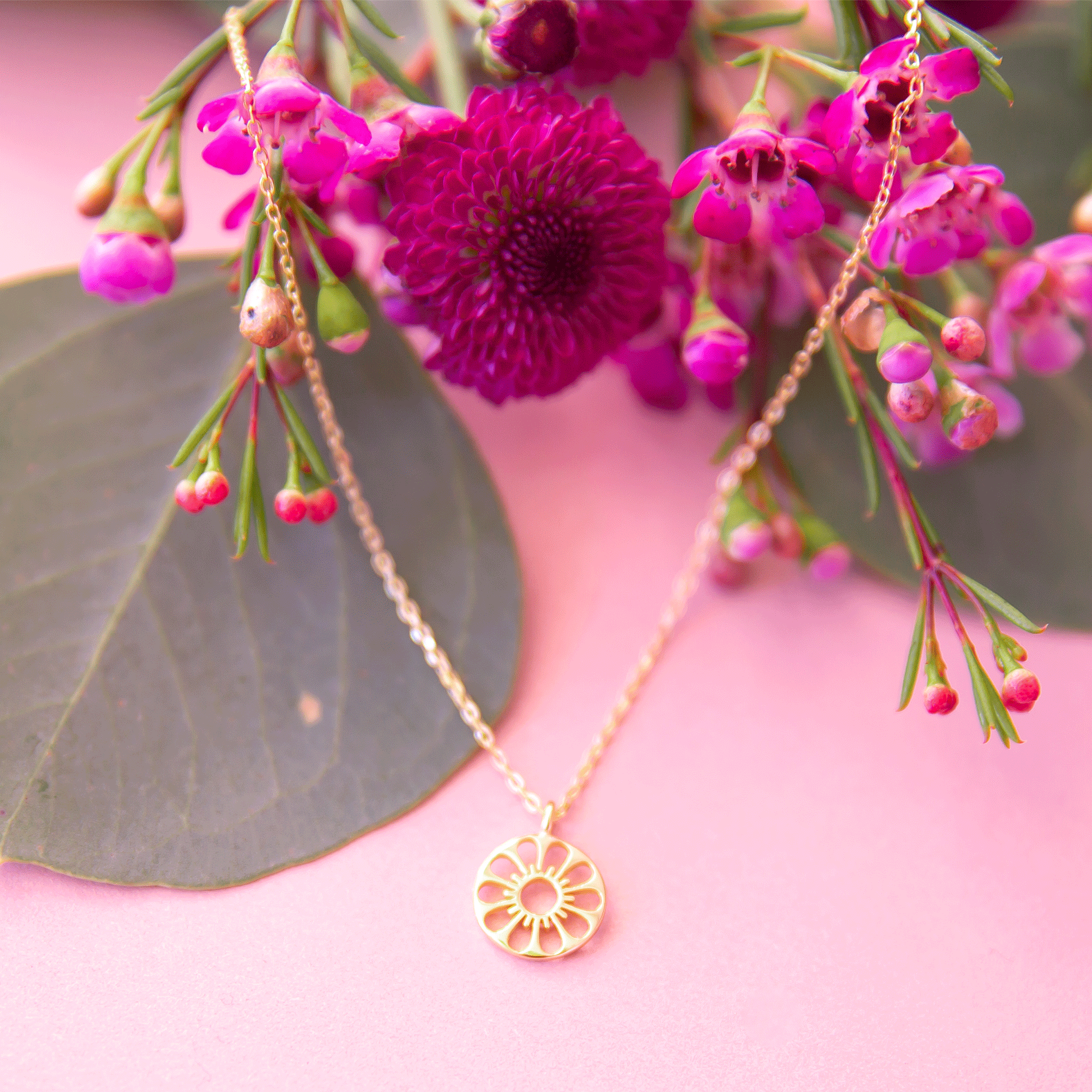 A dainty gold chain necklace with a gold circle pendant in the center with a cut out of a daisy flower, photographed next to pink dried florals.