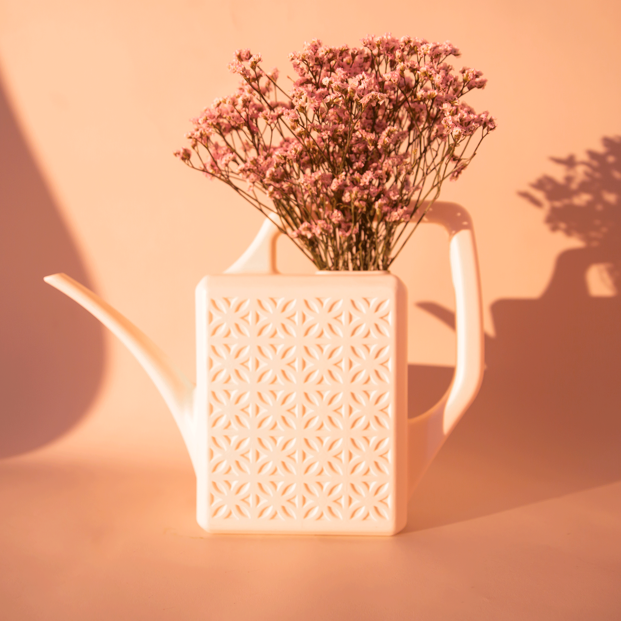 On a peachy background is a white plastic watering can with a long curved spout and a squared off handle along with a breeze block textured design on the sides of the watering can.