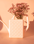 On a peachy background is a white plastic watering can with a long curved spout and a squared off handle along with a breeze block textured design on the sides of the watering can.