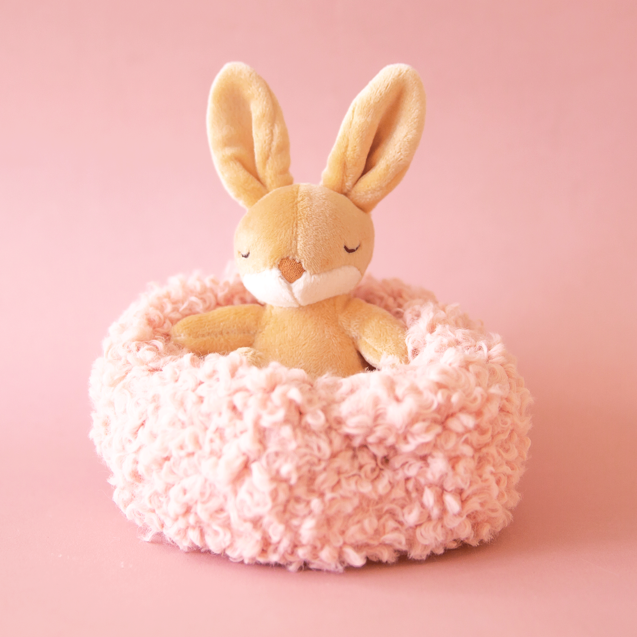 On a pink background is a small bunny stuffed animal in a tan shade sitting inside of a light pink / tan fuzzy nest. 