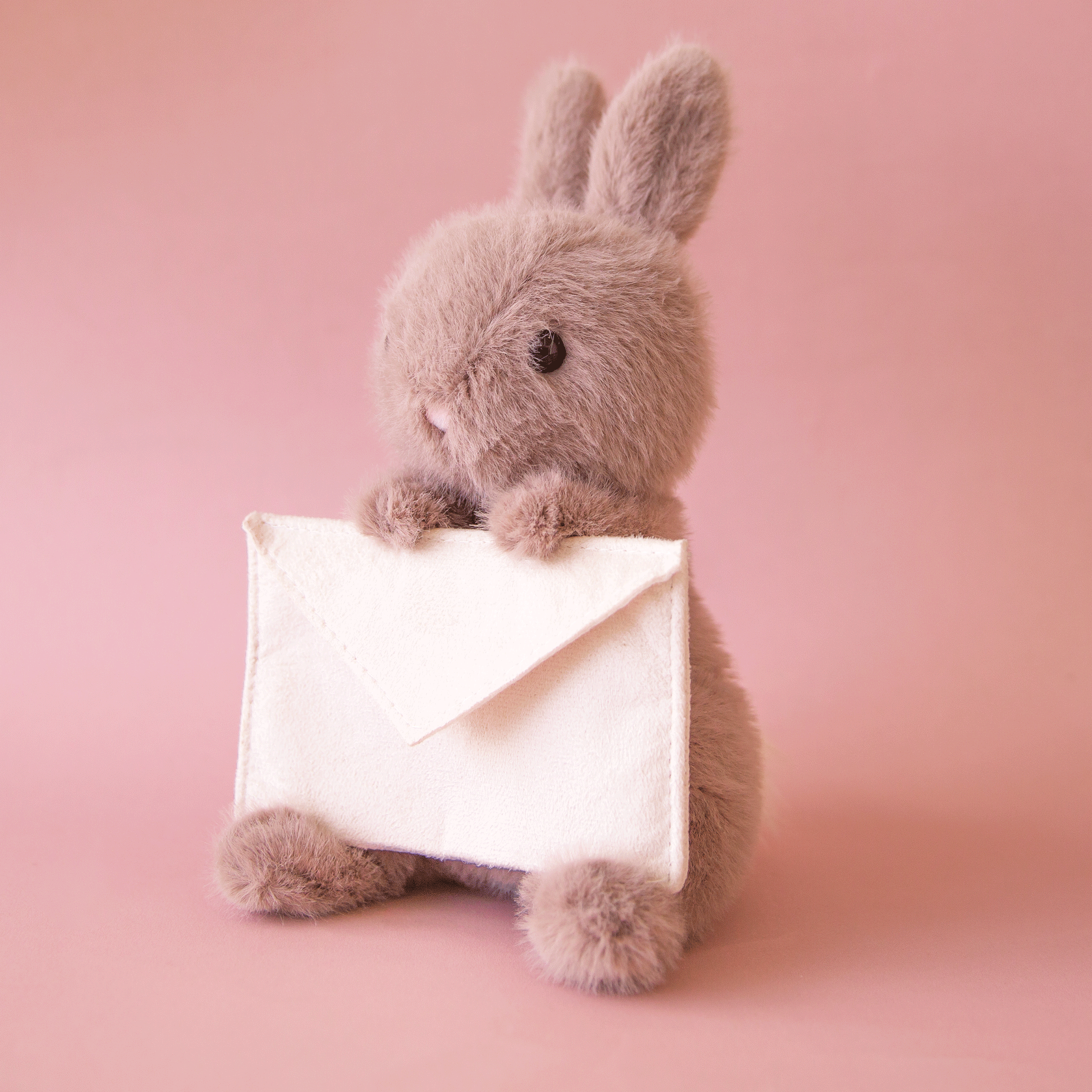 On a pink background is a grey bunny stuffed animal holding an ivory felt envelope. 