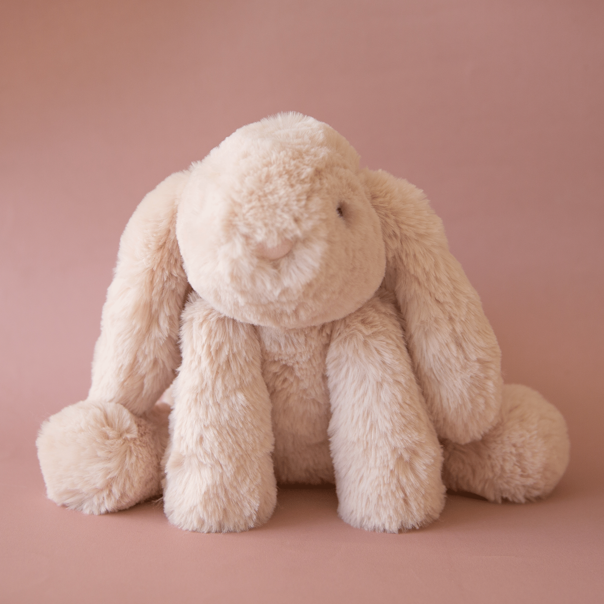 On a pink background is a tan / ivory bunny stuffed animal with floppy ears and a fluffy tail.