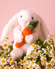 On pink background is a white stuffed toy bunny with long floppy ears and holding an orange stuffed toy carrot.