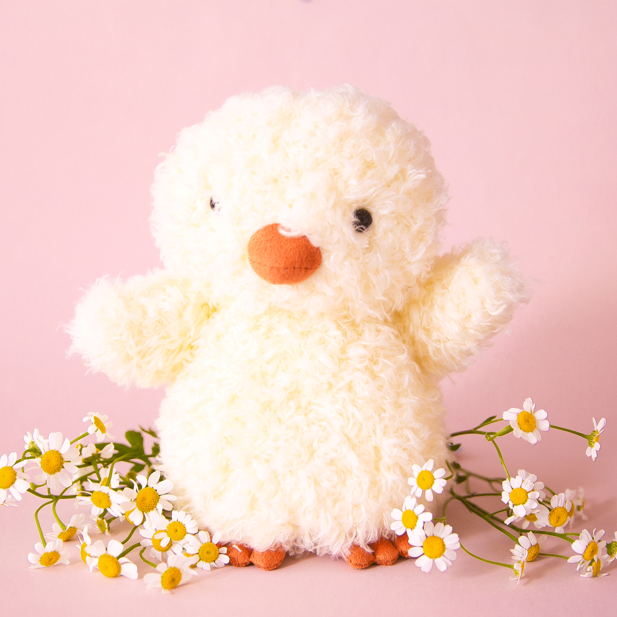 On a pink background is a super light yellow fuzzy chick stuffed animal toy with an orange beak and feet.