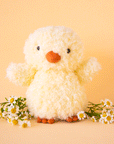 On a yellow background is a super light yellow fuzzy chick stuffed animal toy with an orange beak and feet.
