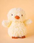 On a yellow background is a super light yellow fuzzy chick stuffed animal toy with an orange beak and feet.
