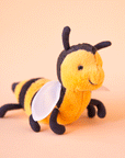 On a peachy background is a yellow and black bee stuffed animal with a smiling face, a fuzzy body and round wings.
