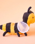 On a peachy background is a yellow and black bee stuffed animal with a smiling face, a fuzzy body and round wings.