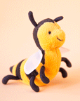 On a peachy background is a yellow and black bee stuffed animal with a smiling face, a fuzzy body and round wings.