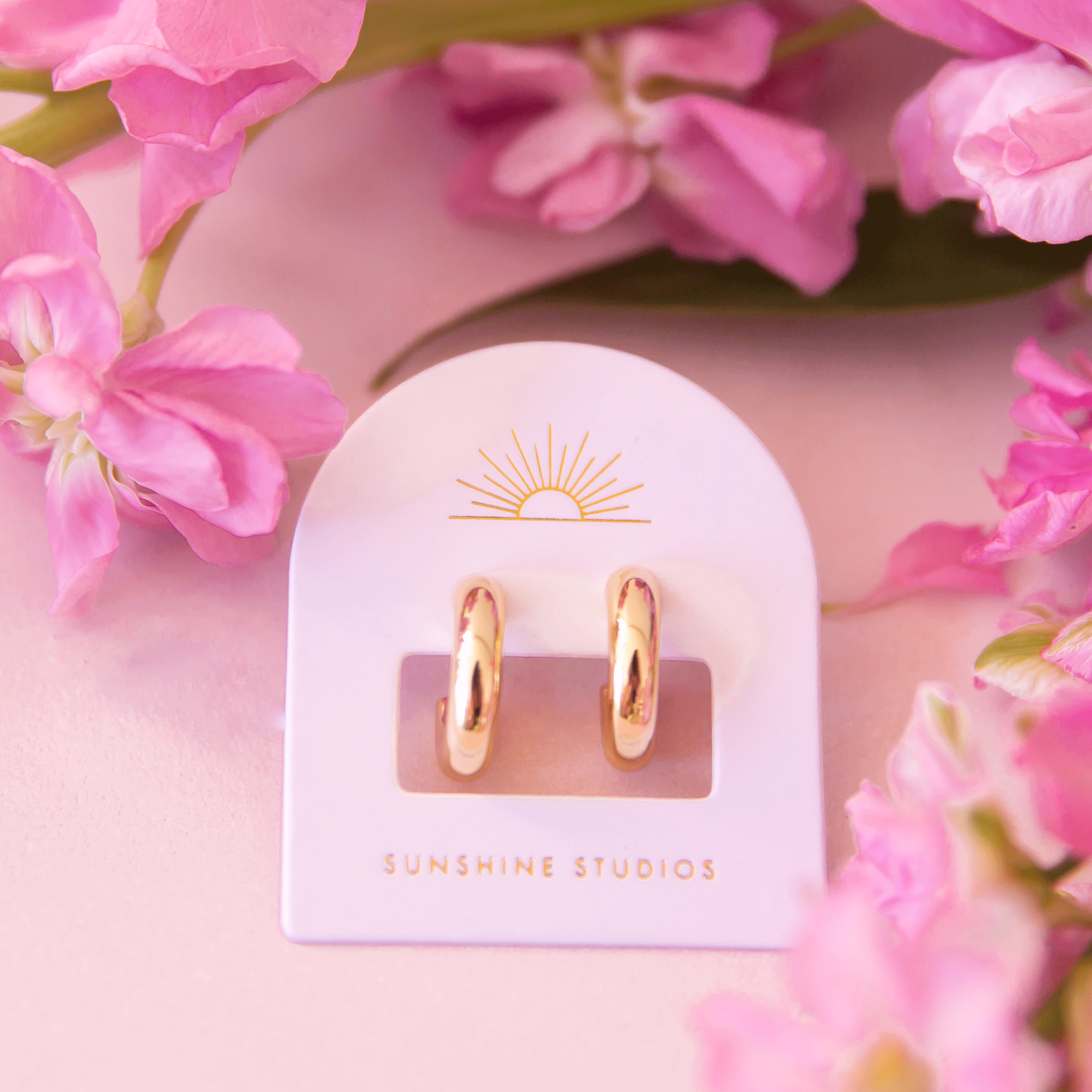 On a pink background is a pair of chunky gold hoop earrings on their arched packaging with a sun ray logo at the top and gold text at the bottom that reads, &quot;Sunshine Studios&quot;.