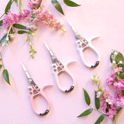 On a pink background is all three color ways of the pruning shears next to bouquets of flowers.