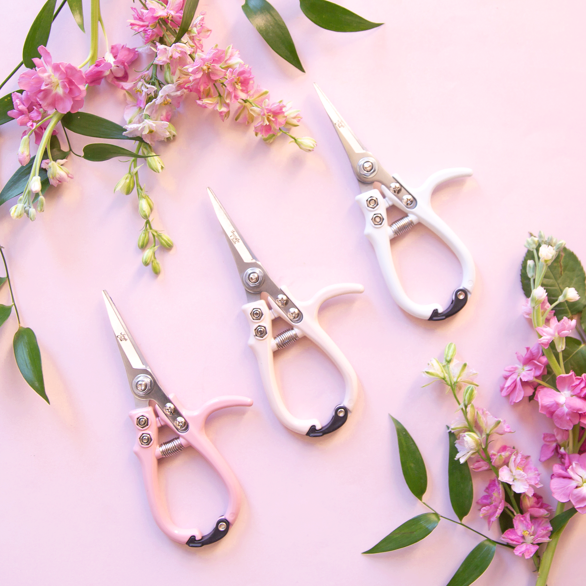 On a pink background is the three color ways of pruning shears next to bouquets of flowers.