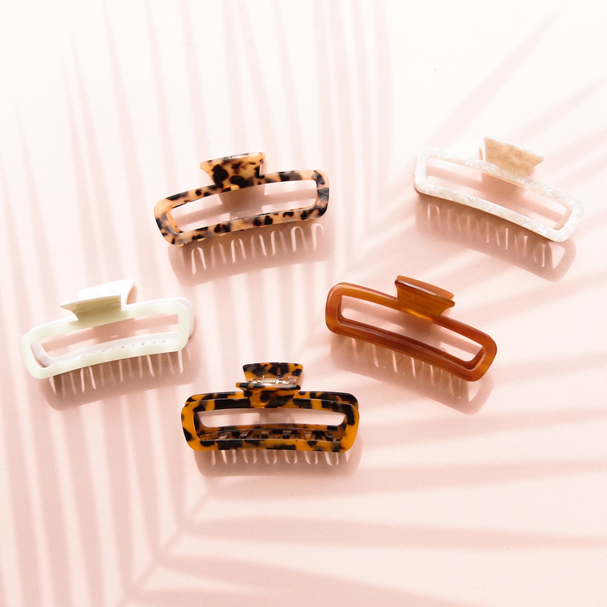 On a light pink background is a variety of rectangle claw clips in tortoise shades, an amber color and two different whites.