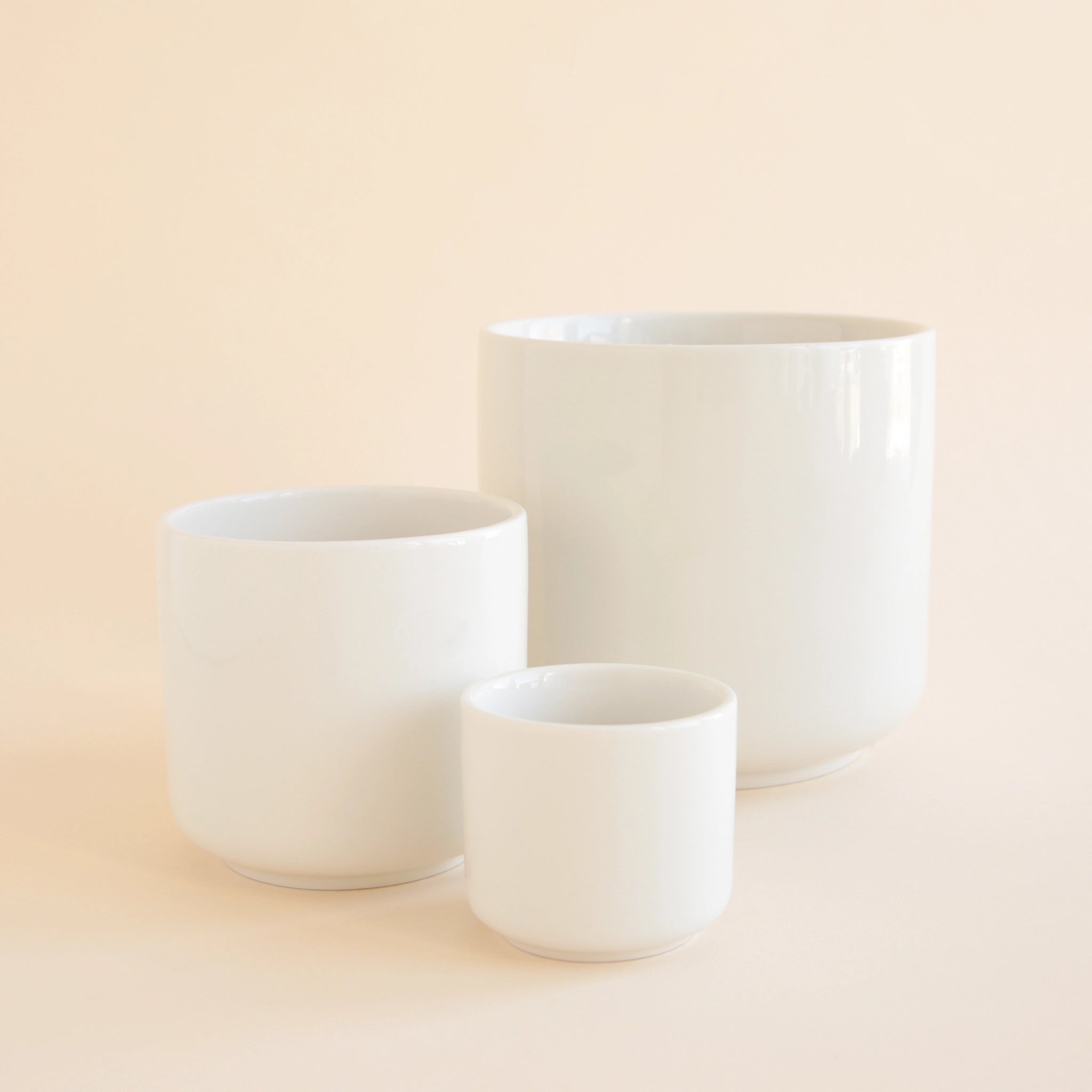 On a tan background is three different sized white ceramic planters with a glossy finish.