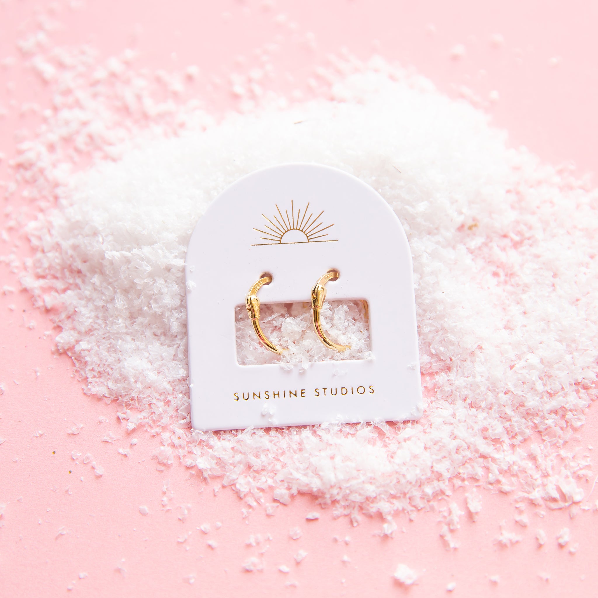 On a pink snowy background is a pair of thin gold hoop earrings on their arched packaging that features a sun ray logo at the top and gold text at the bottom that reads, &quot;Sunshine Studios&quot;.
