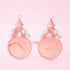 On a pink background is a pair of holiday glasses with tree shapes and six white pom poms and a star on top.