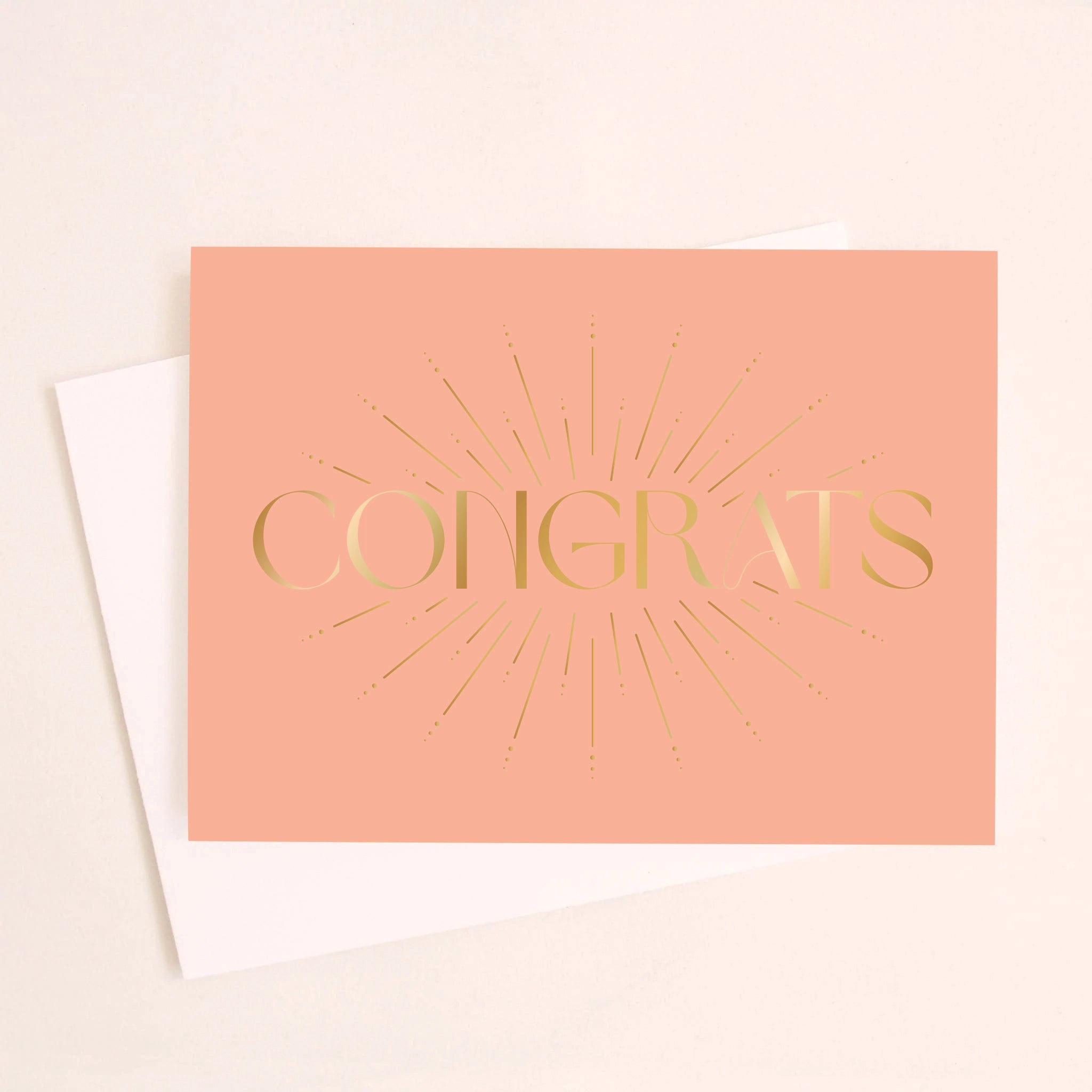 On an ivory background is a salmon colored greeting card with gold foil text that reads, &quot;Congrats&quot; along with a white envelope.