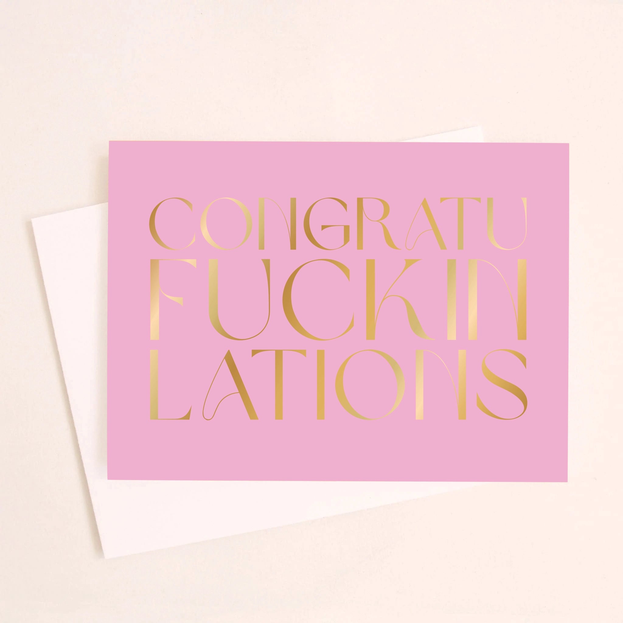 On an ivory background is a purple pink greeting card with gold foil text that reads, &quot;Congraufuckinlations&quot;.