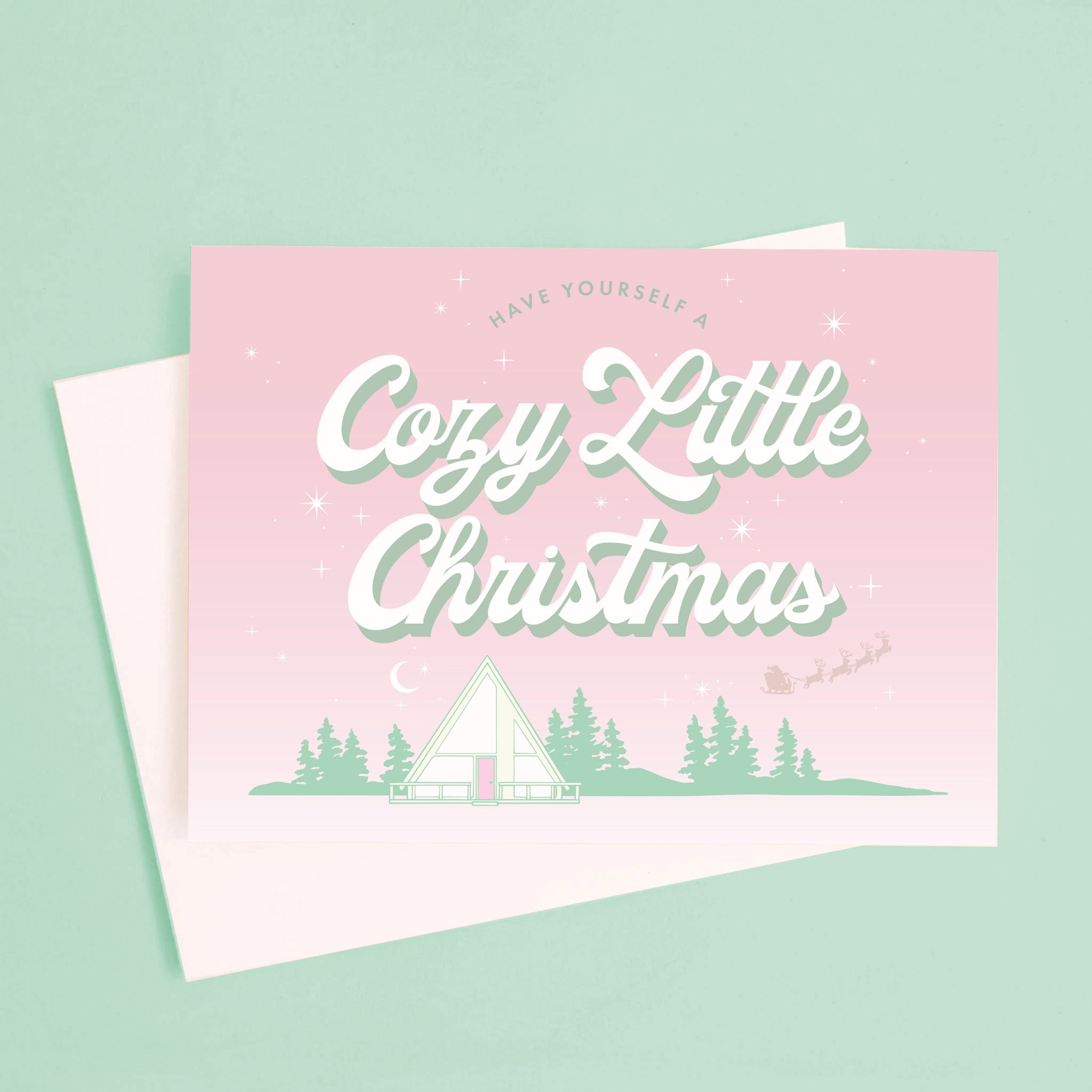 A pink and mint green Christmas card with an A-frame graphic and text above that reads, "Have Yourself A Cozy Little Christmas".