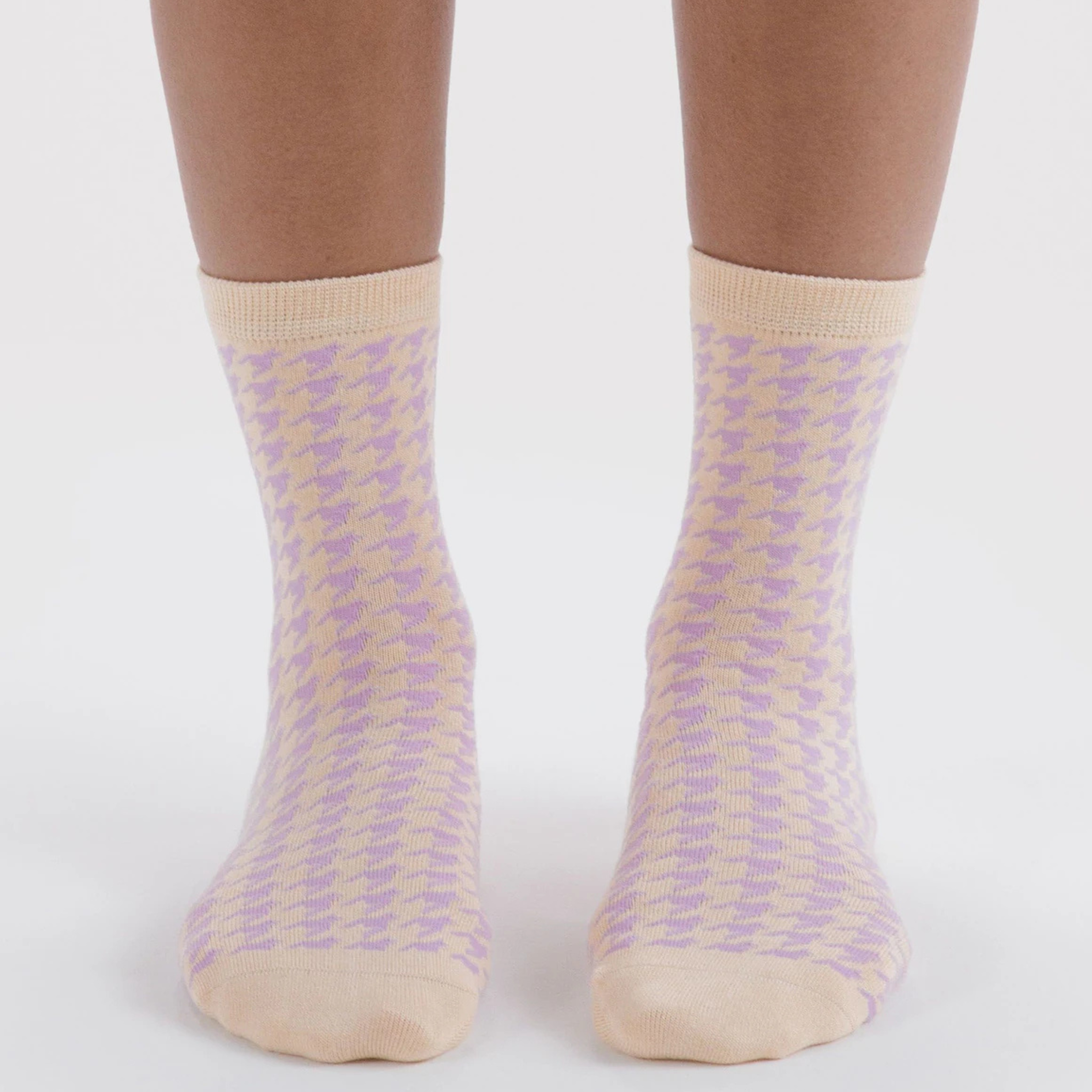 A pair of light cream crew socks with a subtle lavender houndstooth pattern and olive green 'BAGGU' branding printed on the sole, displayed on a warm beige background.
