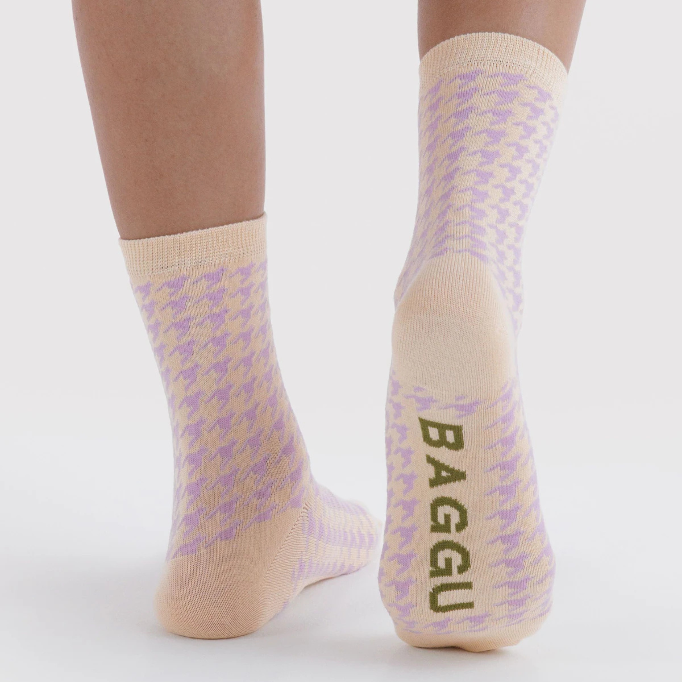 A pair of light cream crew socks with a subtle lavender houndstooth pattern and olive green 'BAGGU' branding printed on the sole, displayed on a warm beige background.
