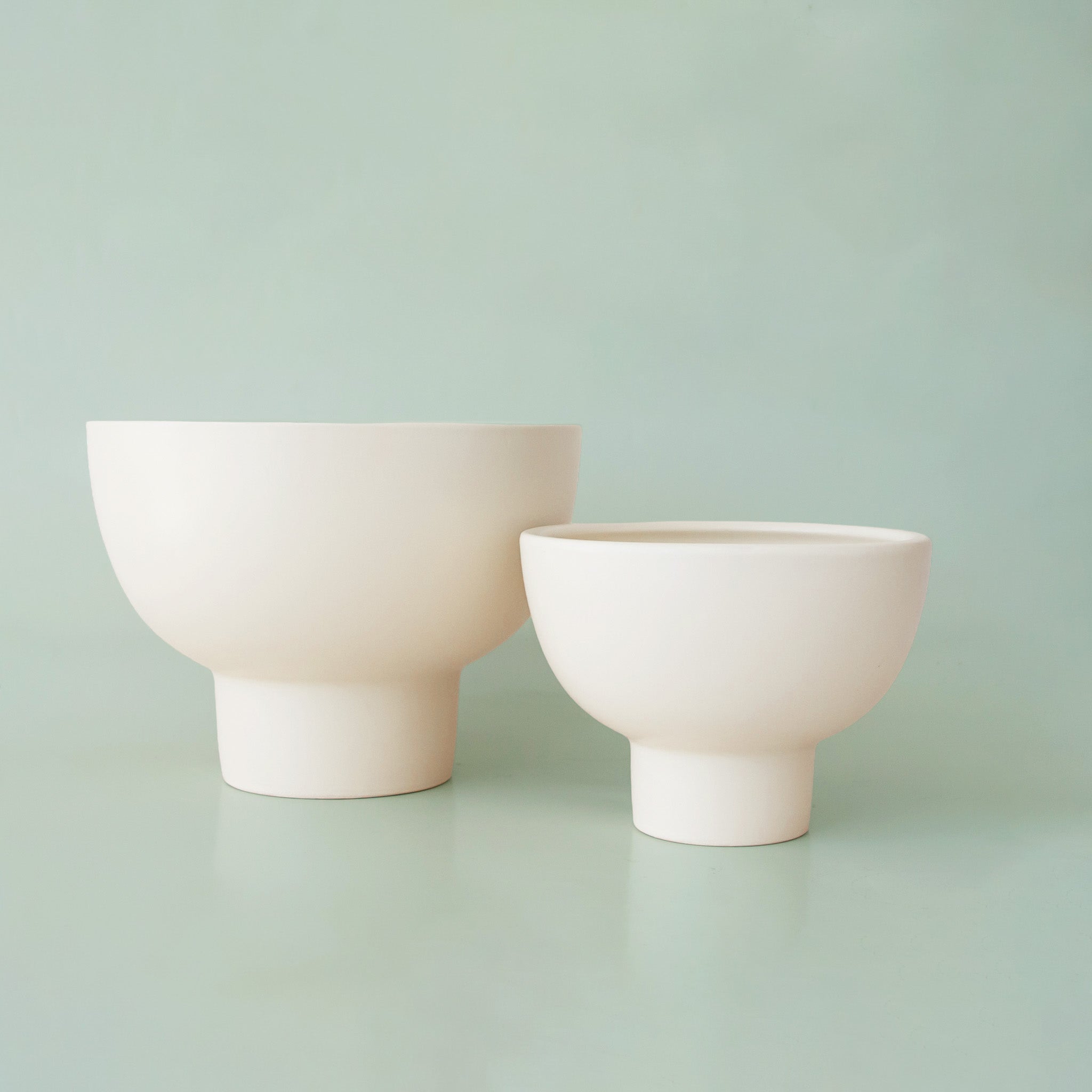 Two ceramic pedestal planters on a light green background