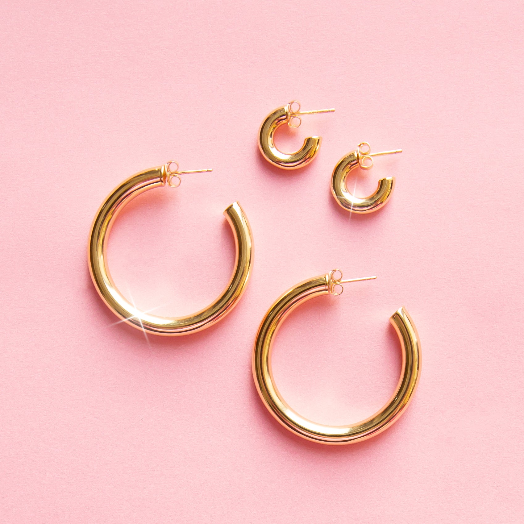 On a pink background is a three different sized pairs of chunky gold hoop earrings with a straight post backing. Each pair sold separately. 