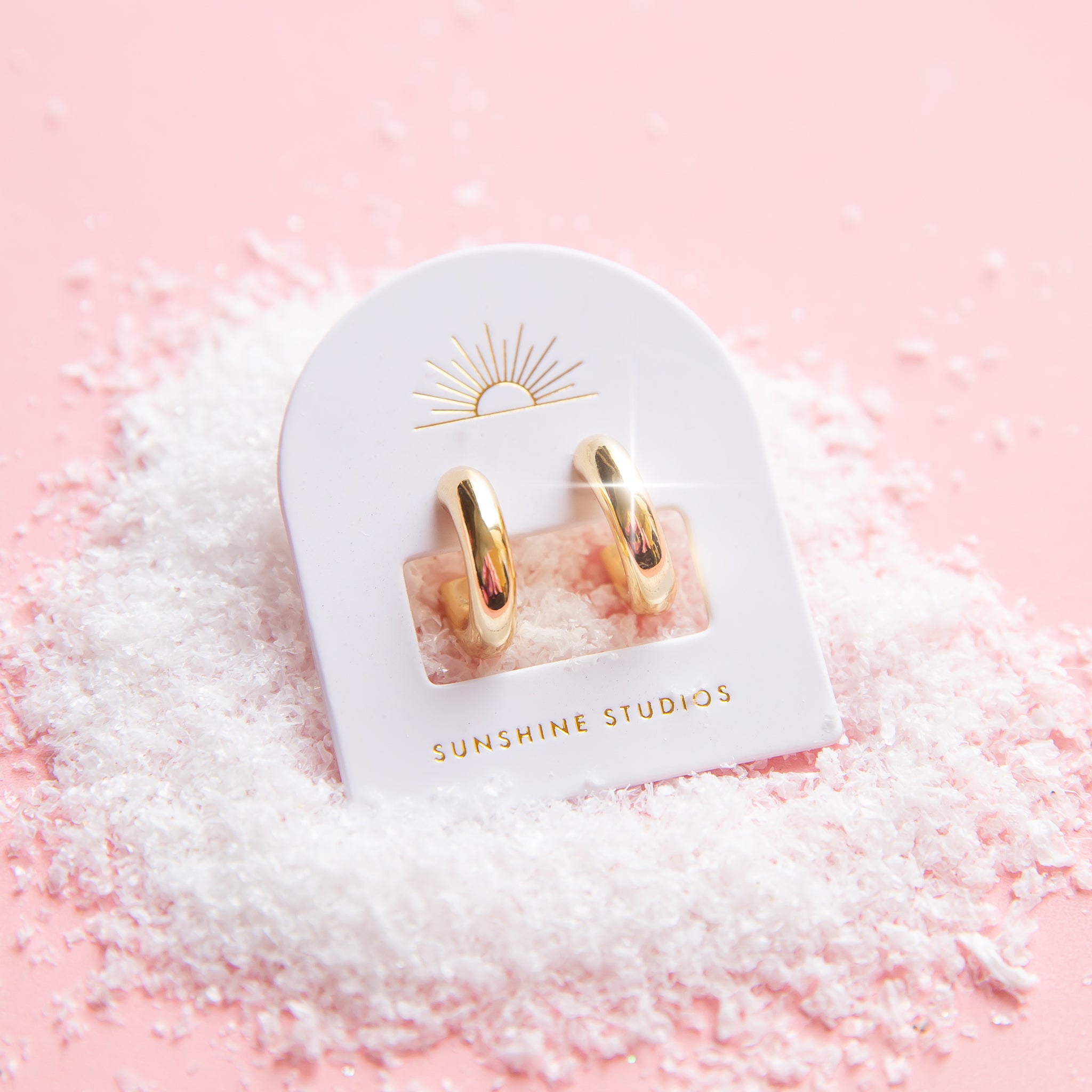 On a snowy pink background is a pair of chunky gold hoop earrings on their arched packaging with a sun ray logo at the top and gold text at the bottom that reads, &quot;Sunshine Studios&quot;.