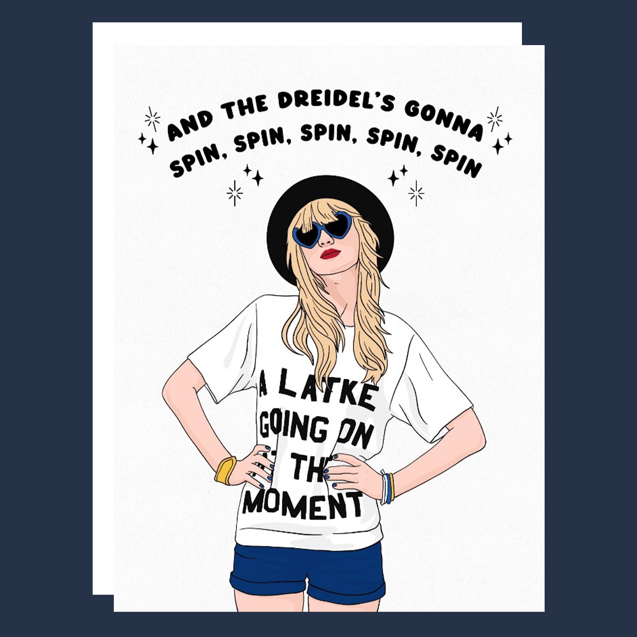 A greeting card sits on a light blue background. The white card with white envelope behind feature an illustration of Taylor Swift standing with her hands on her hips wearing jean shorts and a white t-shirt that reads "A Latke Going on at the Moment".  Above her head the card reads in black font, "And the dreidel's gonna spin, spin, spin, spin, spin". 
