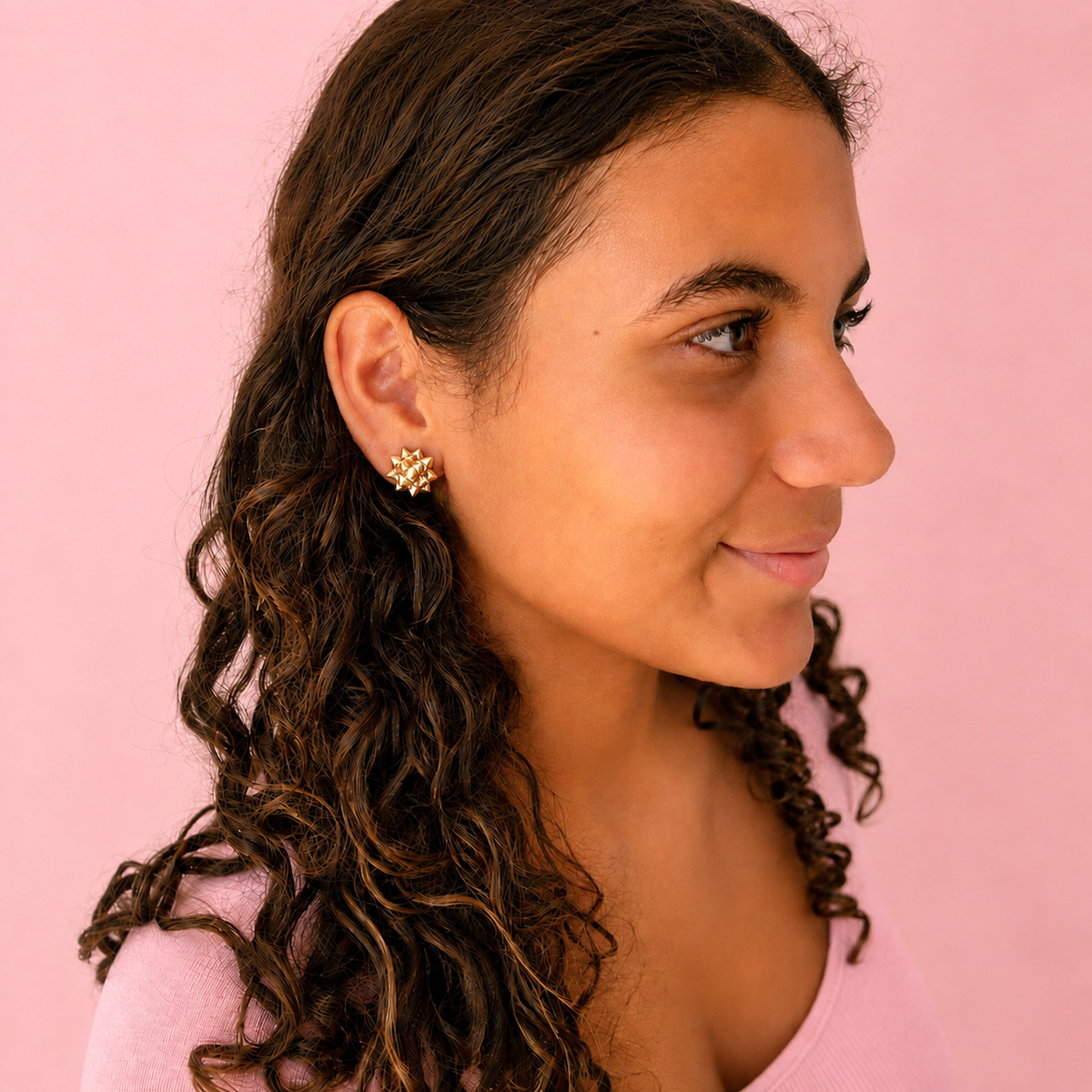 A model wearing a pink top wears gold bow shaped earrings.