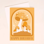 On an ivory background is an orange card with an arched design and illustrations of mushrooms and a small snail on top of the largest one along with text on the bottom that reads, "Happy Birthday". 