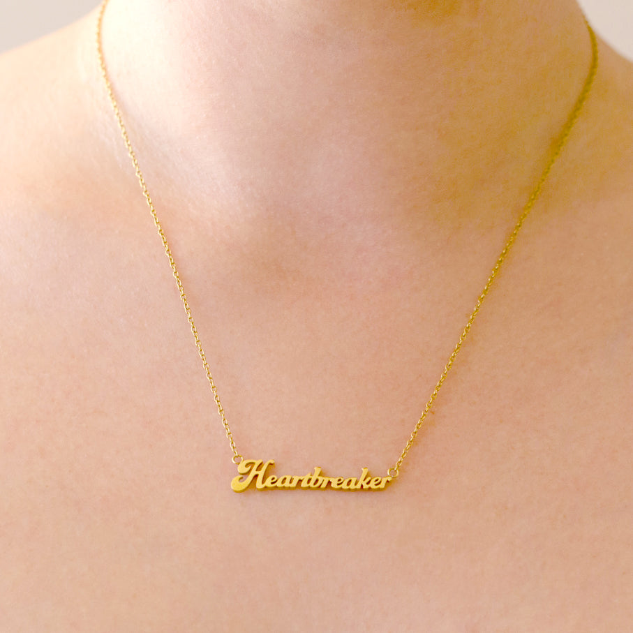 A gold name plate style necklace that reads, "Heartbreaker". 