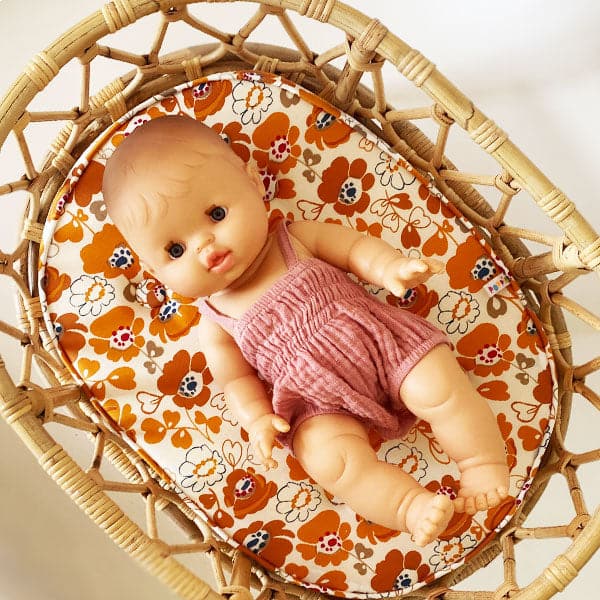 A European baby girl doll wearing a pink romper jumpsuit and laying in a rattan crib with a floral pattern cushion.