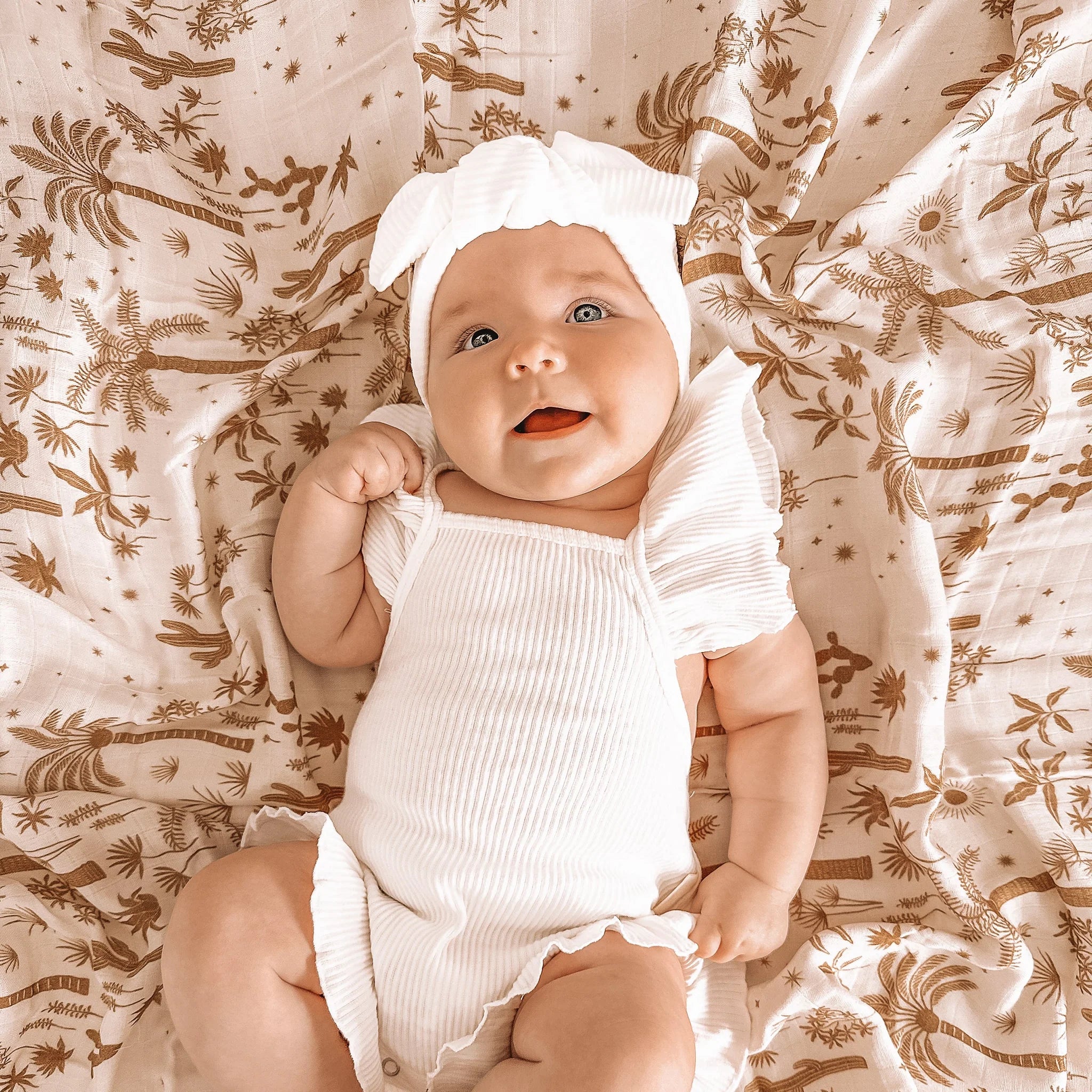 A baby laying on a light pink / neutral swaddle with a light brown palm and desert pattern swaddle. 