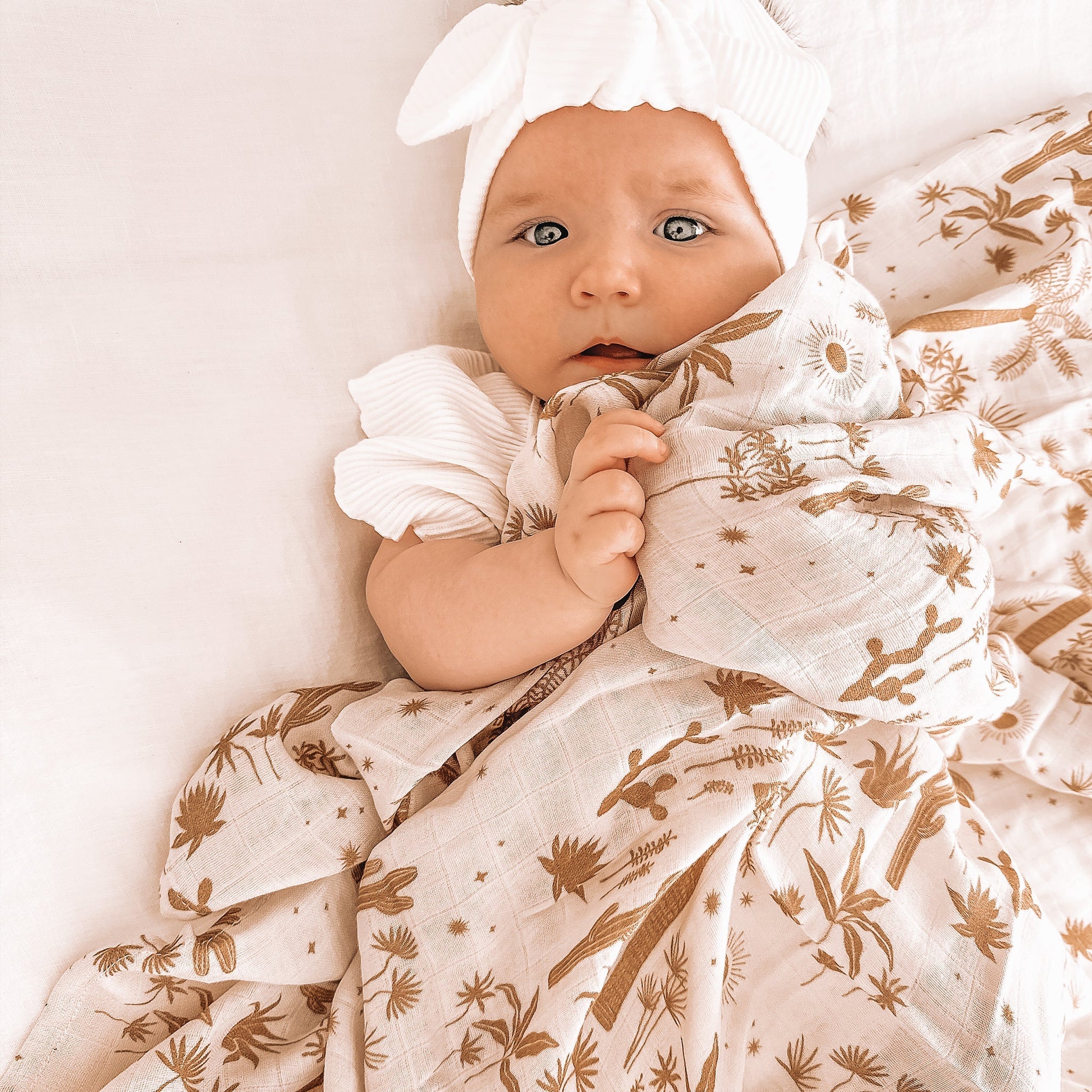 A baby laying on a light pink / neutral swaddle with a light brown palm and desert pattern swaddle.