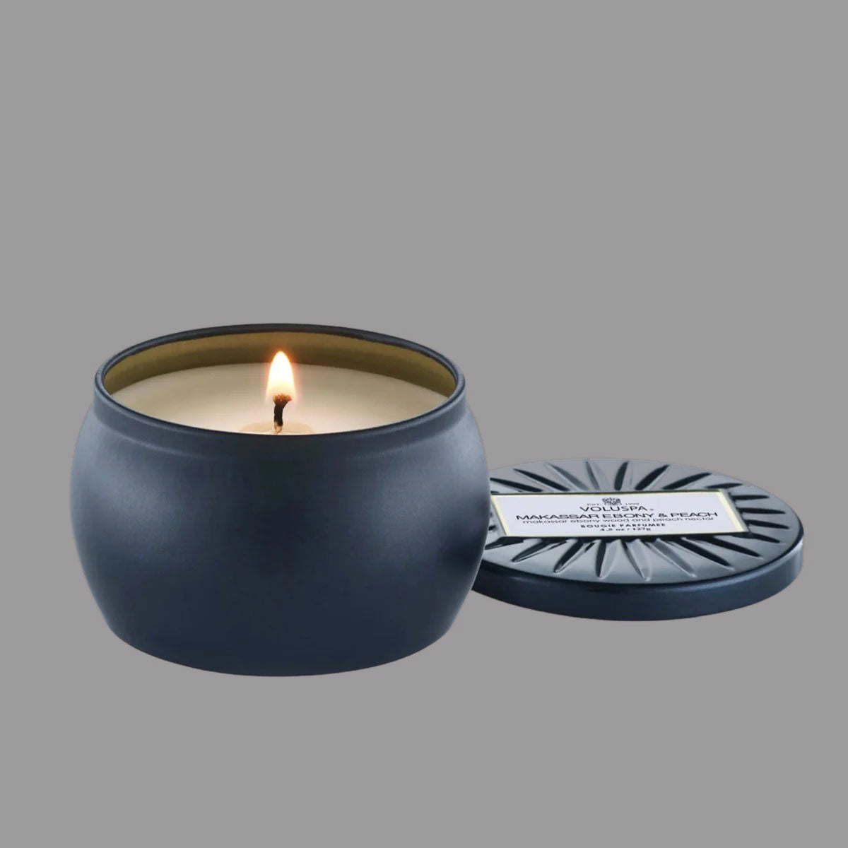 On a cream background is a round tin single wick candle in a navy blue shade along with a label on the lid that reads, "Voluspa Makassar Ebony & Peach".