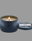 On a cream background is a round tin single wick candle in a navy blue shade along with a label on the lid that reads, "Voluspa Makassar Ebony & Peach".