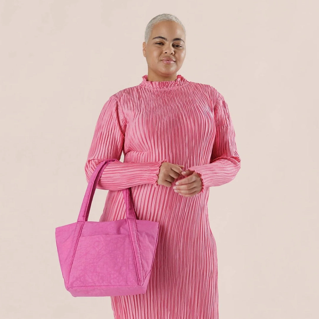 On a tan background is a bright pink tote bag made of durable nylon fabric with two large handles for over the shoulder and an outside pocket.