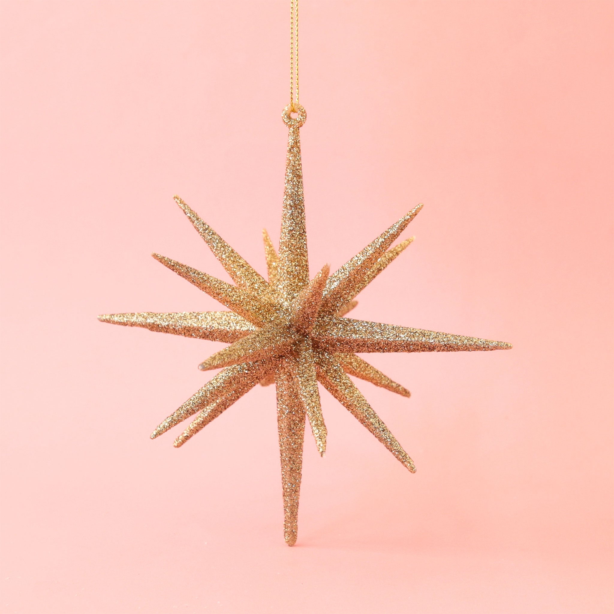 On a peachy pink background is a gold glitter 16 point startburst ornament. 