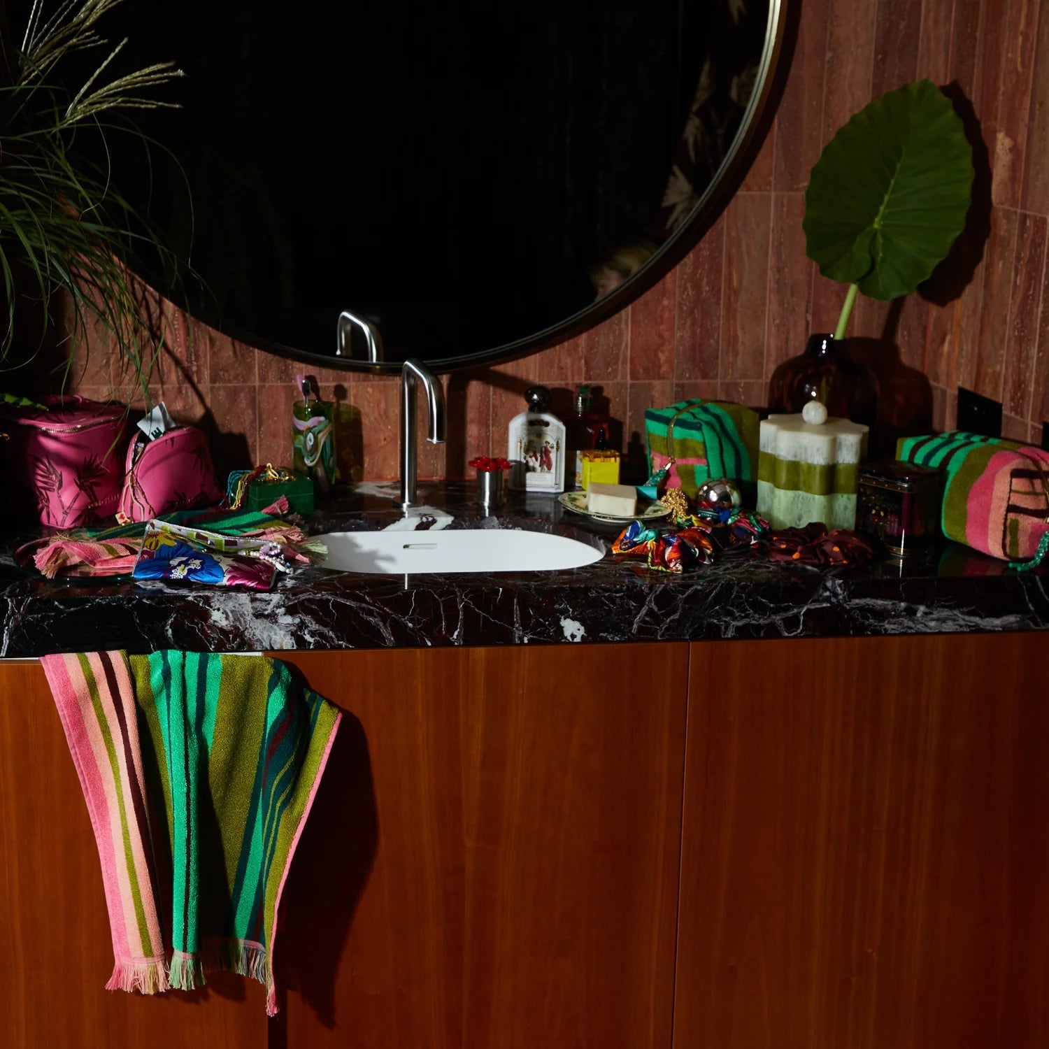 Styled bathroom vanity with a striped hand towel in pink, green, and olive draped over a wooden cabinet, surrounded by colorful accessories and a round mirror above.