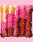 On a pink background is a multi colored shag pillow in orange, brown, pink and yellow shades. 