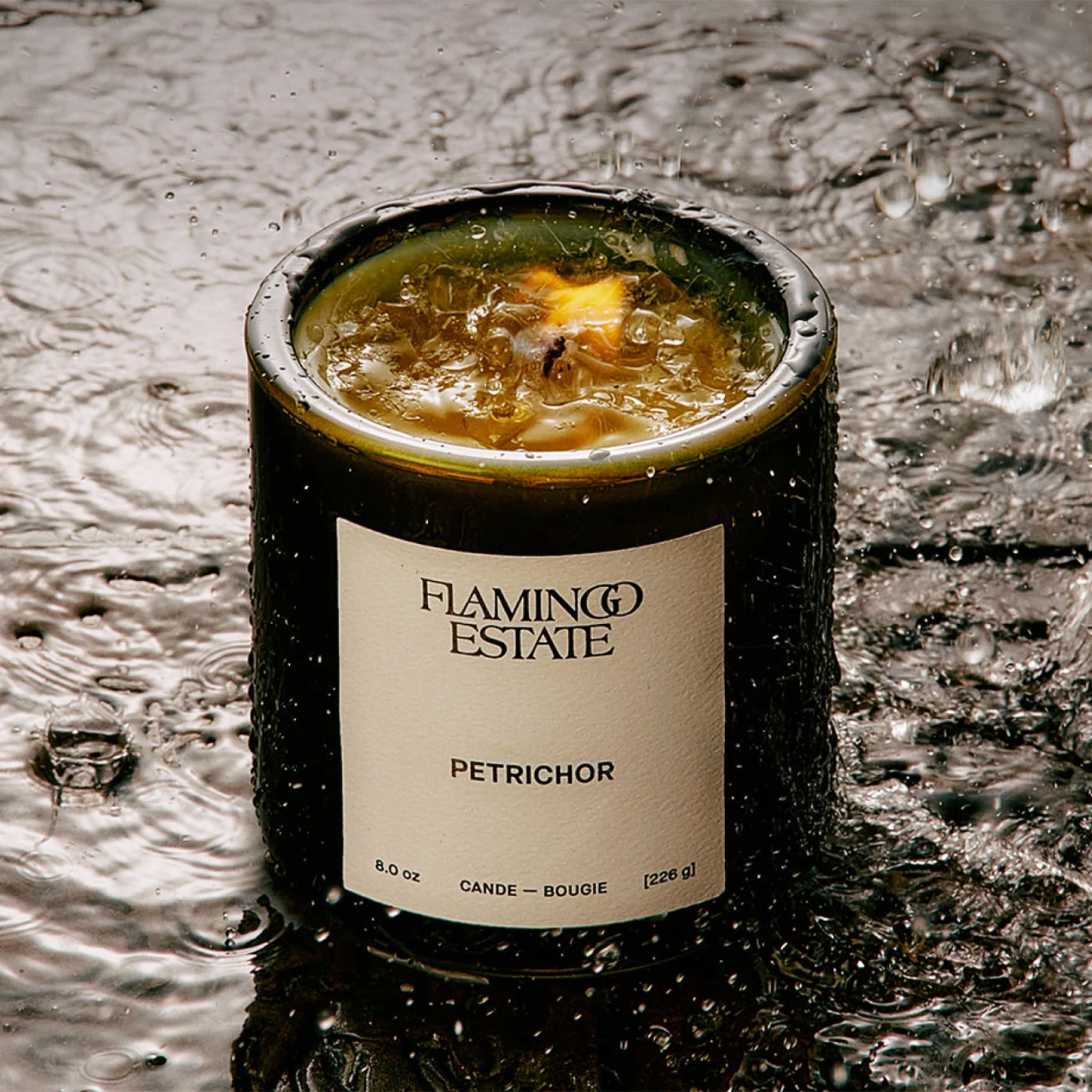 A green glass candle with a white label with text that reads, 'Flamingo Estate petrichor'. 
