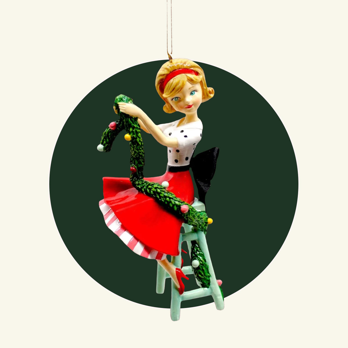 A resin ornament of a retro style girl wearing a red skirt and polka dot top.  She is holding green Christmas decorating garland and sitting on a mint stool.