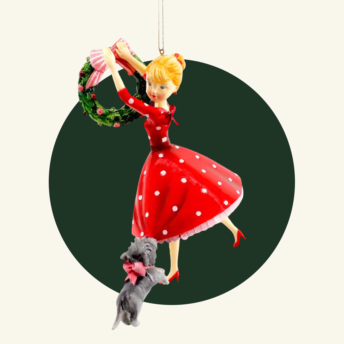 A retro looking girl wearing a big red dress with polka dots.  A gray dog tugs at her dress while she hangs a Christmas wreath.