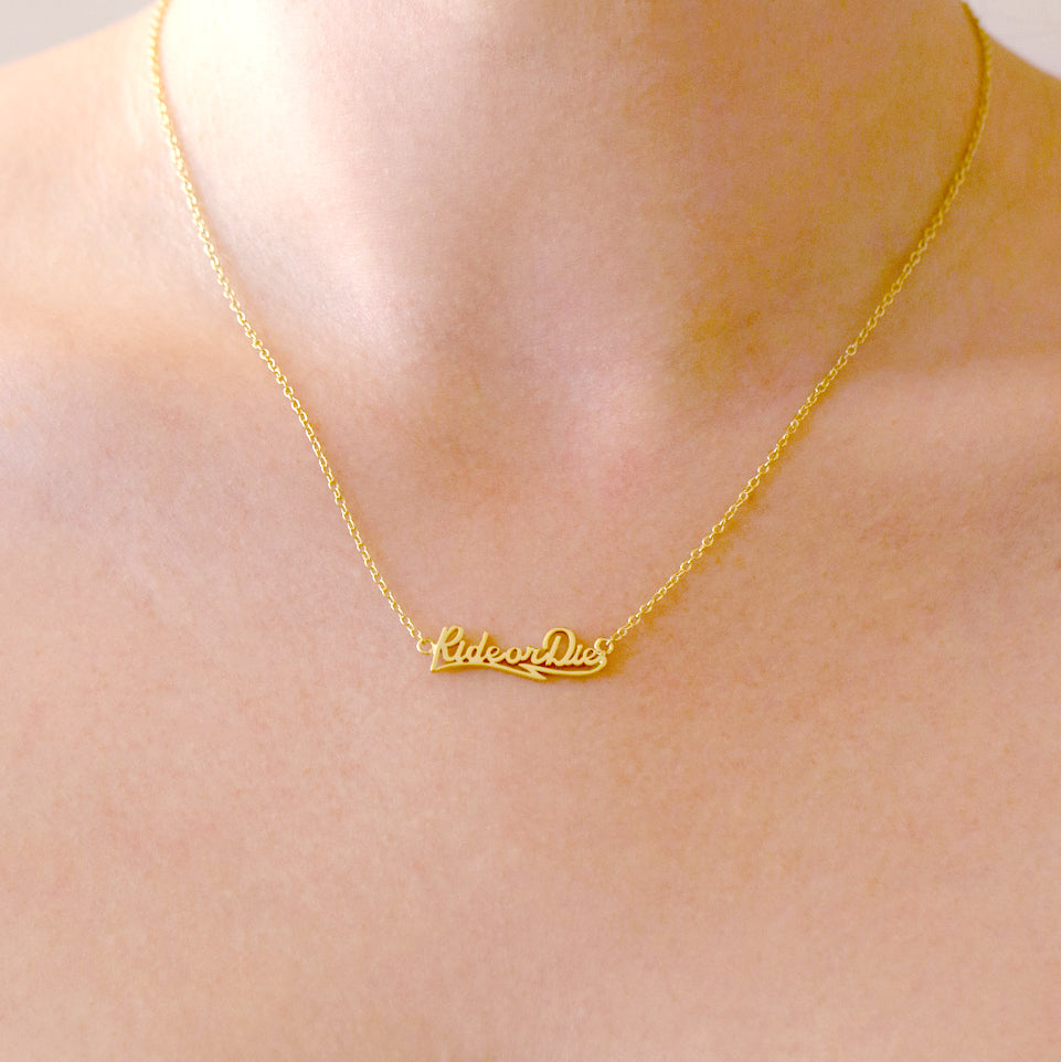 A gold nameplate style necklace with text that reads, "RideorDie". 