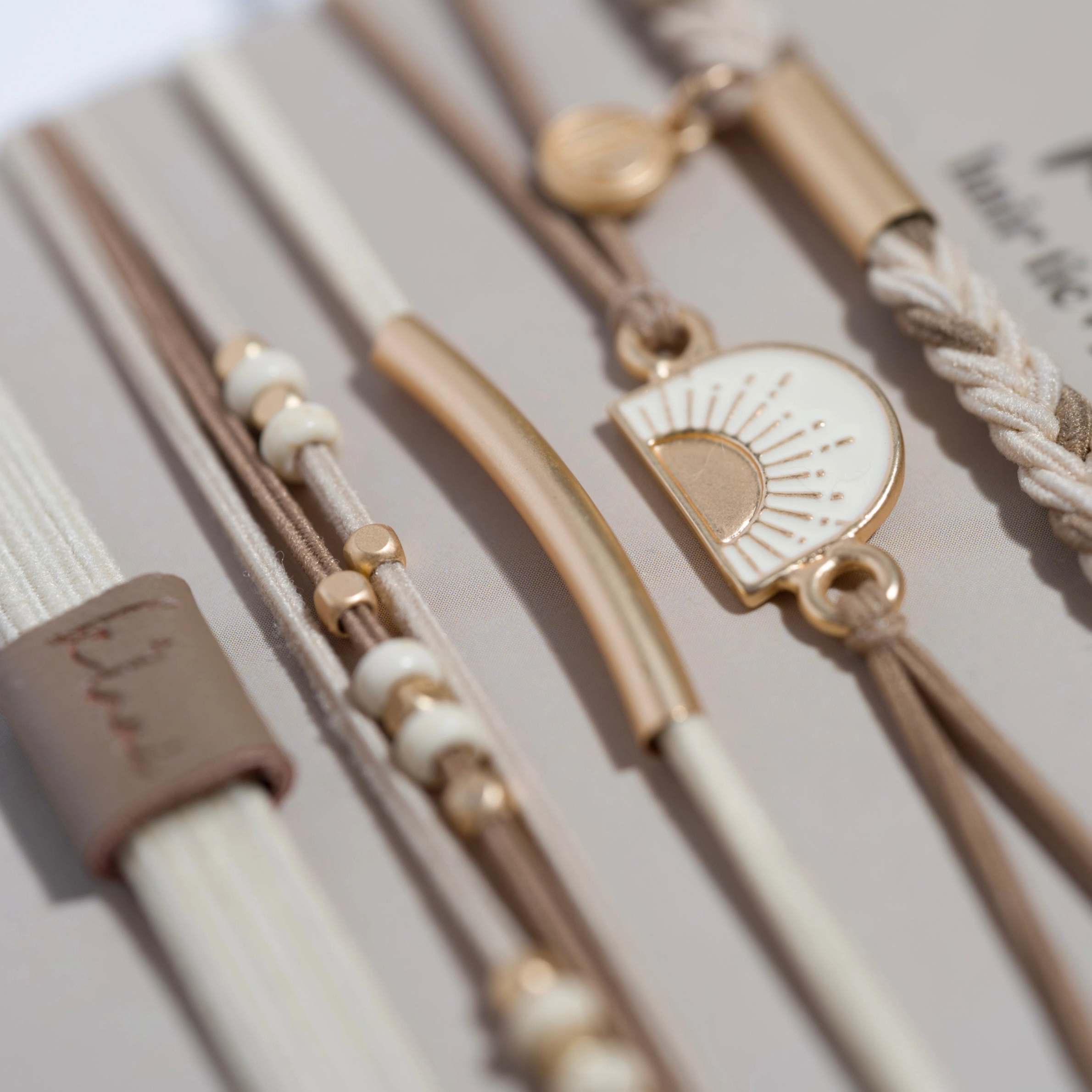 A set of five bohemian-style hair ties and bracelets displayed on a beige backing card. The set includes braided elastic bands, thin brown cords, a gold-tone sun charm, and small bead accents.
