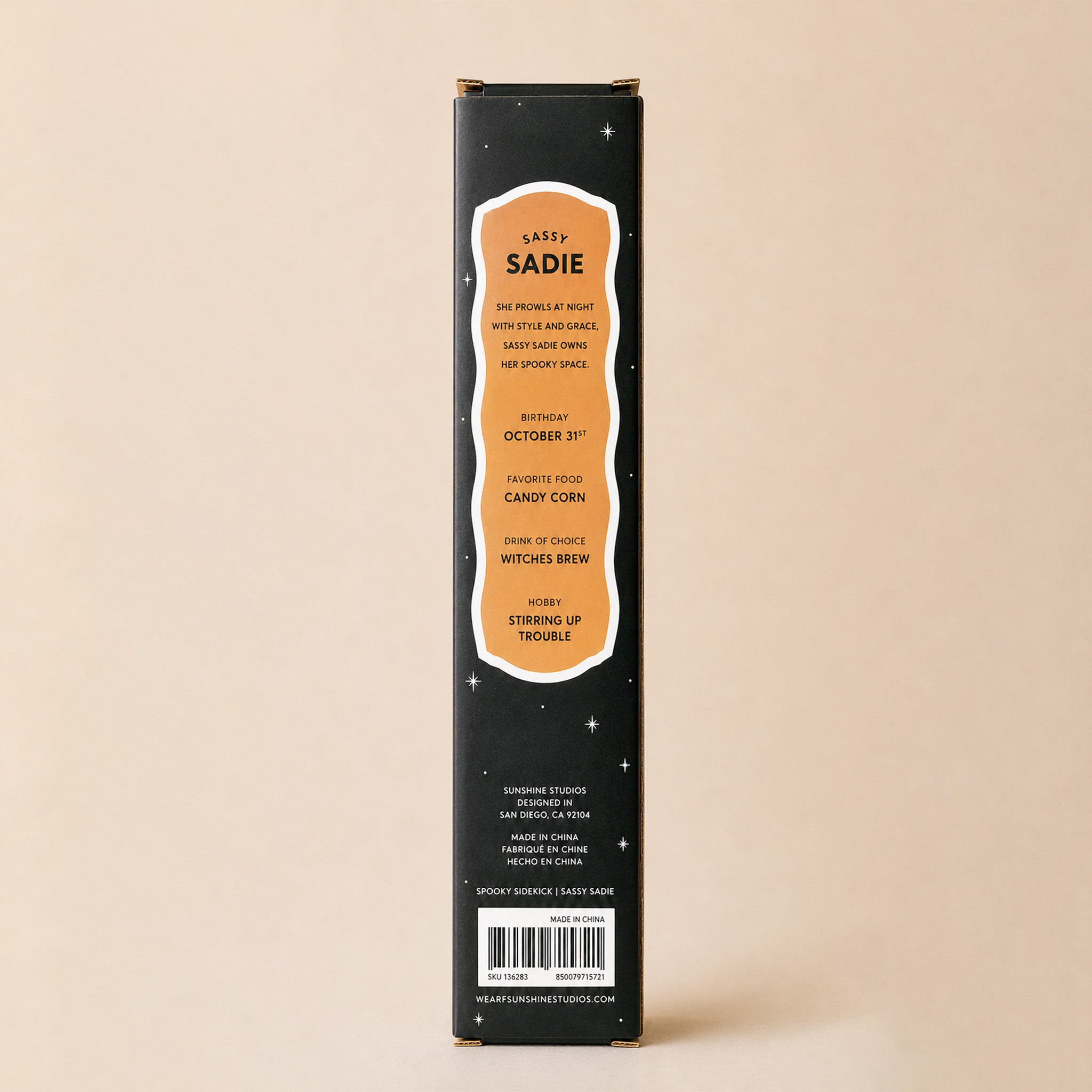 A tall, narrow black product box standing upright against a warm beige background. The front has a large peach label with white text reading 'SADIE' and additional details, plus a barcode near the bottom.