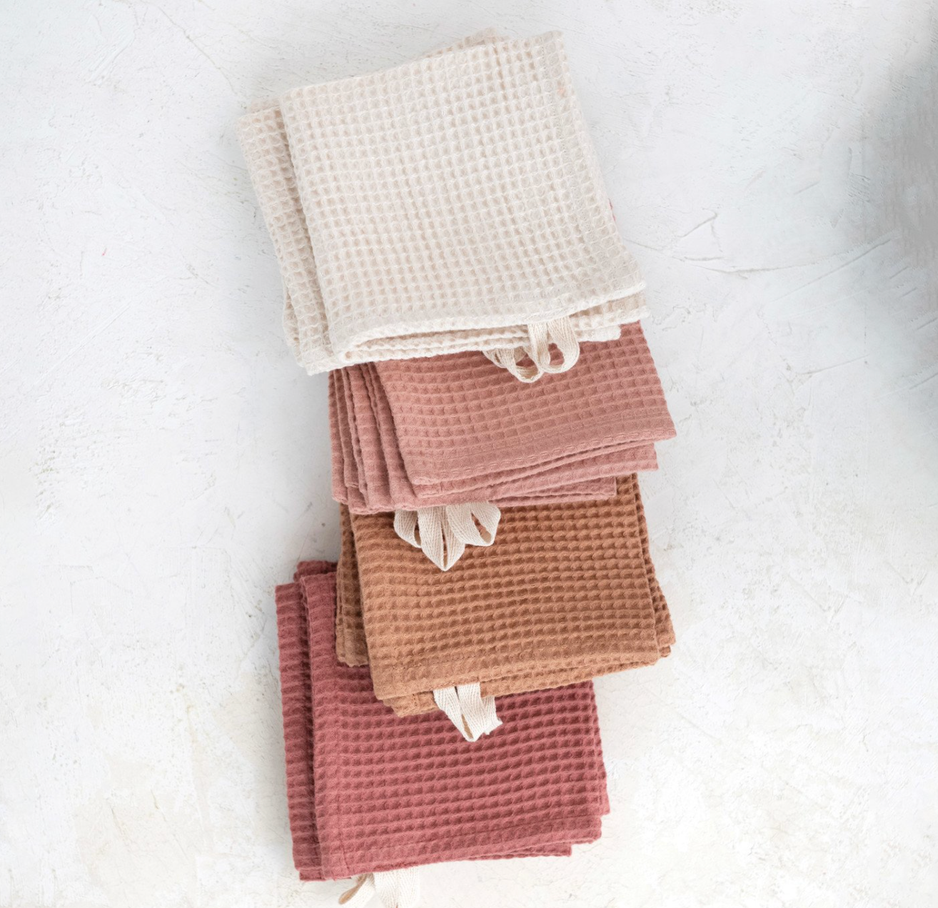 On a white background is four linen cotton waffle knit dish clothes with loops on the end for hanging. There are four colors. From top to bottom there is ivory, blush, terracotta and rose. 