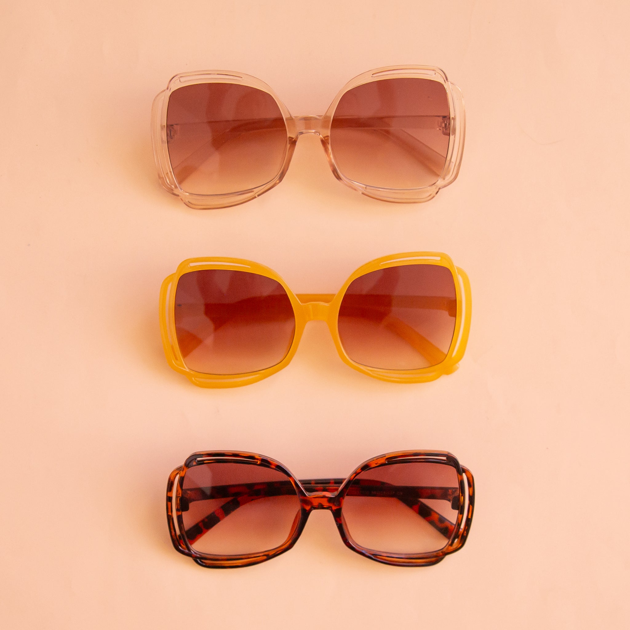 On a peachy background is a pair of tortoise sunglasses with a round frame and a brownish pink lens sitting next to the other two available color options.