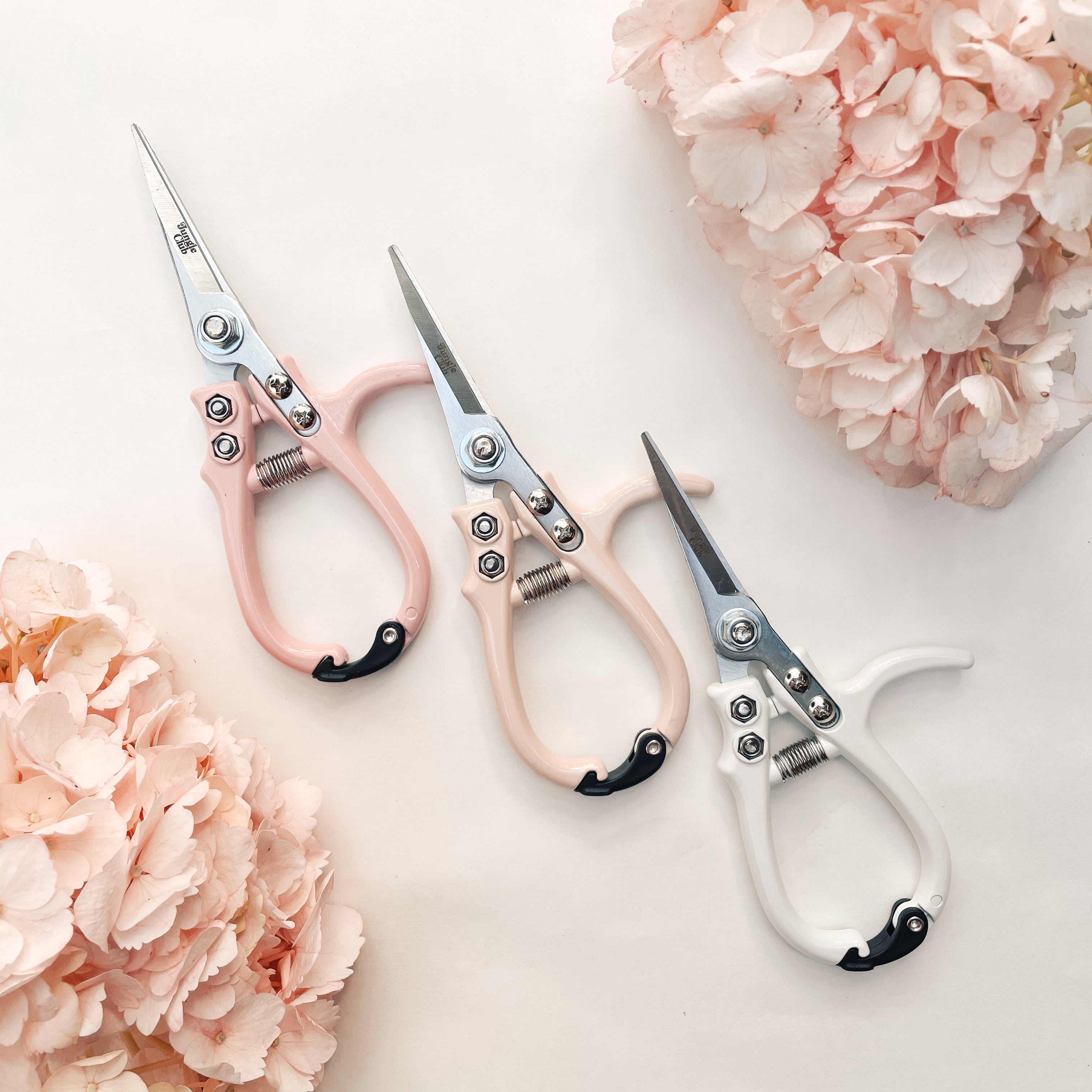 Pruning shears with a narrow tip, and light cream colored handle and a black clasp to connect when shears are not in use. There is small black text on the side of the shears that read, "Jungle Club".